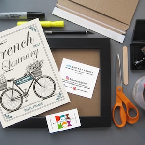May include: A white framed print with a black and white illustration of a bicycle with a basket full of flowers and the text "French Laundry Daily". The print is on a gray surface with other crafting supplies, including a black frame, scissors, tape, and a stamp.