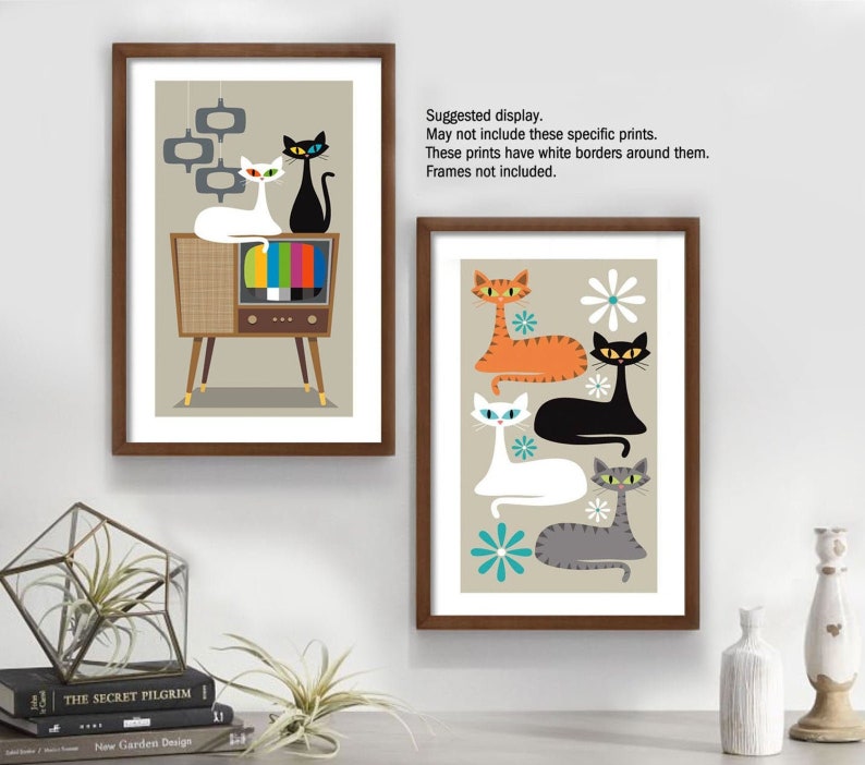 May include: Two framed prints featuring retro-style illustrations of cats. The first print shows three cats in front of a vintage television set. The second print shows four cats with a floral background. Both prints have a beige background.