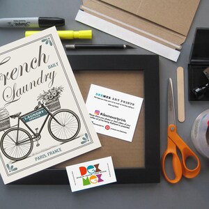 Puede incluir: Una impresi&oacute;n enmarcada en blanco con una ilustraci&oacute;n en blanco y negro de una bicicleta con una cesta llena de flores y el texto "French Laundry Daily". La impresi&oacute;n est&aacute; sobre una superficie gris con otros materiales de artesan&iacute;a, incluyendo un marco negro, tijeras, cinta adhesiva y un sello.