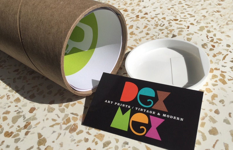 May include: A brown cardboard tube with a green and white logo. The tube is open and a white plastic lid is next to it. A black business card with the text "Dex Mex Art Prints / Vintage & Modern" is in the foreground.