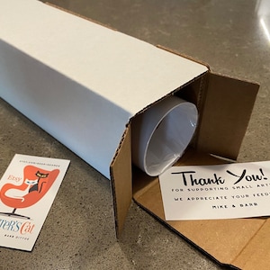 May include: A white cardboard tube with a thank you note inside. The note says "Thank You! For supporting small artists. We appreciate your feedback. Mike & Barb". The tube is sitting on a gray surface. There is a business card for "Dexter's Cat" on the left side of the image.