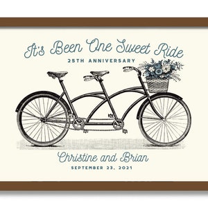 May include: A black tandem bicycle with a basket of flowers on the handlebars. The text "It's Been One Sweet Ride" is above the bicycle, and "25th Anniversary" is below. The names "Christine and Brian" and the date "September 23, 2021" are below the bicycle.