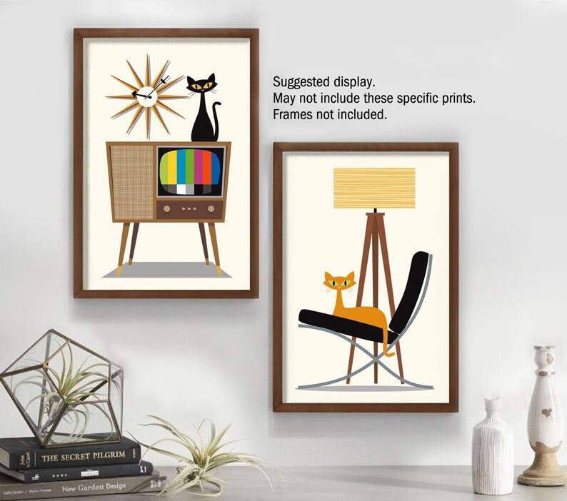 May include: Two framed prints with a retro mid-century modern theme. The first print features a black cat sitting on top of a vintage television with a clock behind it. The second print features an orange cat sitting in a black chair with a tripod lamp behind it.  "Suggested display. May not include these specific prints. Frames not included."