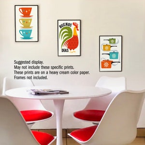 Op de afbeelding: Drie ingelijste prints met een mid-century modern esthetiek. De prints tonen een haan met de tekst "Buenos Dias", een stapel theekopjes en een stapel potten. De prints worden getoond op een witte muur boven een witte tafel met rode stoelen.