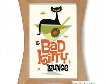 May include: A framed print featuring a black cat perched on a martini glass with an olive and a swizzle stick. The text "The Bad Kitty Lounge" is in orange and black. The frame is made of light-colored wood.