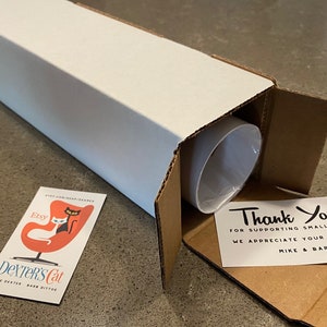 K&ouml;nnte beinhalten: Ein wei&szlig;er Karton mit einer Dankeskarte im Inneren. Die Karte lautet "Vielen Dank! F&uuml;r die Unterst&uuml;tzung kleiner K&uuml;nstler. Wir sch&auml;tzen Ihr Feedback. Mike & Barb". Der Karton ist ge&ouml;ffnet und eine wei&szlig;e R&ouml;hre befindet sich im Inneren. Eine Visitenkarte mit einer schwarzen Katze und einer wei&szlig;en Katze, die in einem roten Ei-Sessel sitzen, liegt auf dem Tisch. Die Visitenkarte lautet "Etsy Dexter's Cat Mike Dexter Barb Ditto".