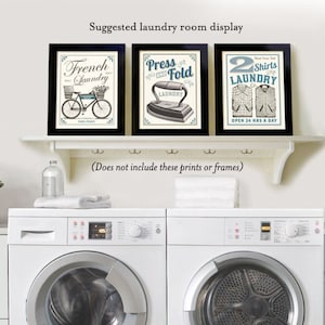 May include: Three framed vintage-style prints with laundry-themed designs. The prints are displayed on a white shelf above two white washing machines. The prints feature a bicycle, an iron, and a shirt.