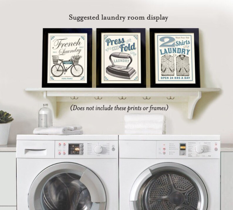 May include: Three framed vintage-style prints with laundry-themed designs. The prints are displayed on a white shelf above two white washing machines. The prints feature a bicycle, an iron, and a shirt.