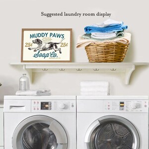 Puede incluir: Un estante blanco con una impresi&oacute;n enmarcada marr&oacute;n que dice "Muddy Paws Soap Co. 25&cent;" con una ilustraci&oacute;n en blanco y negro de un perro corriendo. La impresi&oacute;n est&aacute; sobre dos lavadoras blancas.