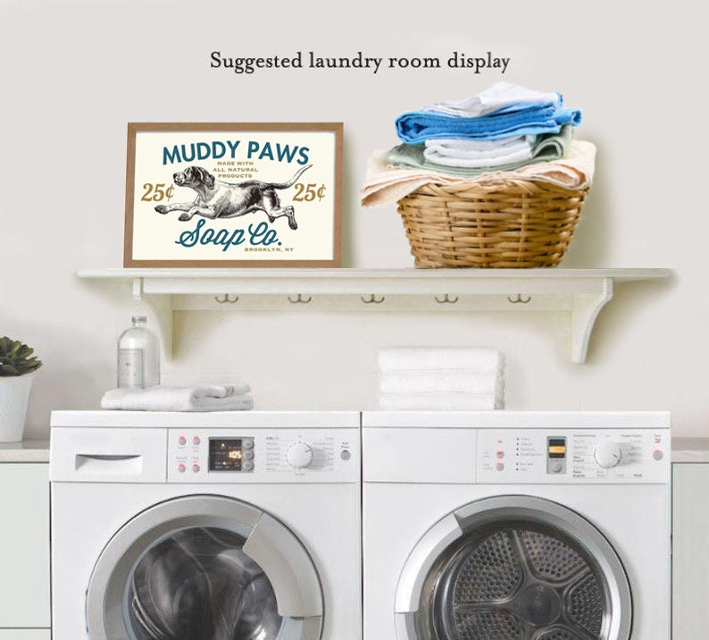 Puede incluir: Un estante blanco con una impresi&oacute;n enmarcada marr&oacute;n que dice "Muddy Paws Soap Co. 25&cent;" con una ilustraci&oacute;n en blanco y negro de un perro corriendo. La impresi&oacute;n est&aacute; sobre dos lavadoras blancas.