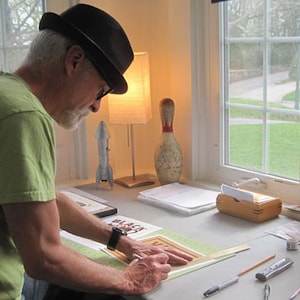 May include: A person wearing a black fedora and a green shirt is working on a craft project at a table. They are using a ruler and a pencil to draw on a piece of paper. There are other craft supplies on the table, including a lamp, a bowling pin, and a rocket model.