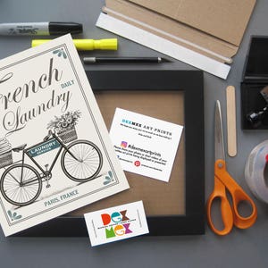 May include: A white and blue print with a black and white illustration of a bicycle with a basket full of flowers. The text on the print reads "French Laundry Daily".