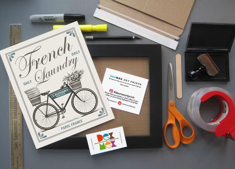 May include: A white and blue print with a black and white illustration of a bicycle with a basket full of flowers. The text on the print reads "French Laundry Daily".