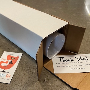 May include: A white cardboard tube with a brown cardboard flap open, revealing a rolled-up print. A thank you note is visible inside the tube, with the text "Thank You! For supporting small artists. We appreciate your feedback. Mike & Barb". A business card with the text "Etsy Dexter's Cat Mike Dexter Barb Dittoe" and a black cat sitting in a red chair is visible on the table.
