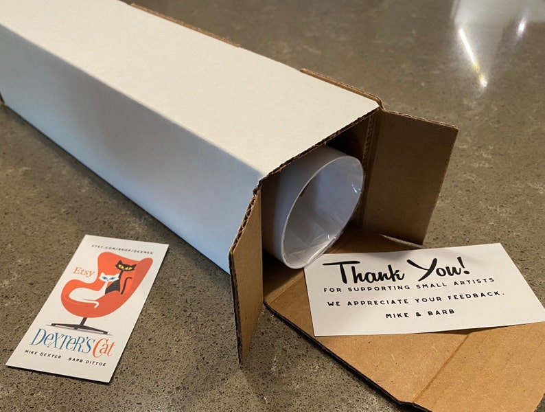 May include: A white cardboard tube with a brown cardboard flap open, revealing a rolled-up print. A thank you note is visible inside the tube, with the text "Thank You! For supporting small artists. We appreciate your feedback. Mike & Barb". A business card with the text "Etsy Dexter's Cat Mike Dexter Barb Dittoe" and a black cat sitting in a red chair is visible on the table.