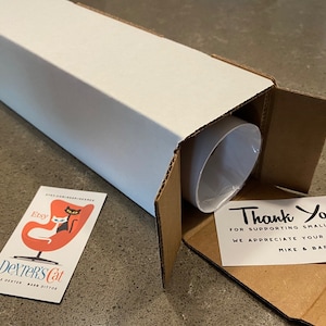 Puede incluir: Un objeto cil&iacute;ndrico blanco es parcialmente visible dentro de una caja de cart&oacute;n blanca. Una peque&ntilde;a tarjeta con un logotipo de gato y el texto "Dexter's Cat" est&aacute; a la izquierda. Una tarjeta de agradecimiento est&aacute; a la derecha, con el texto "Thank You!" y "Mike & Barb."