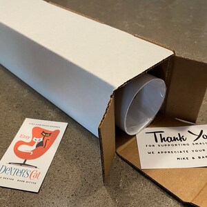 May include: A white cardboard box with a thank you note inside. The note says "Thank You! For supporting small artists. We appreciate your feedback. Mike & Barb". The box is open and a white tube is inside. A business card with a black cat and a white cat sitting in a red egg chair is on the table. The business card says "Etsy Dexter's Cat Mike Dexter Barb Ditto".