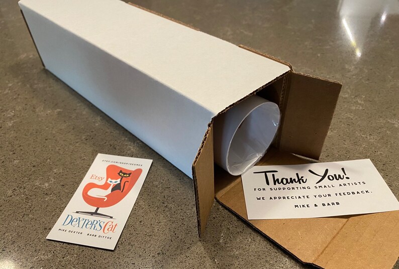May include: A white cardboard box with a thank you note inside. The note says "Thank You! For supporting small artists. We appreciate your feedback. Mike & Barb". The box is open and a white tube is inside. A business card with a black cat and a white cat sitting in a red egg chair is on the table. The business card says "Etsy Dexter's Cat Mike Dexter Barb Ditto".