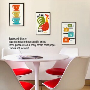 May include: Three framed prints with a mid-century modern aesthetic. The prints feature a rooster with the text "Buenos Dias", a stack of teacups, and a stack of pots. The prints are displayed on a white wall above a white table with red chairs.