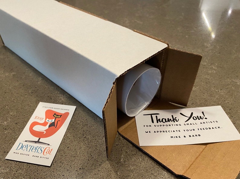 May include: A white cardboard tube with a brown cardboard flap open, revealing a rolled-up print inside. A thank you note is visible inside the tube, with the text "Thank You! For supporting small artists. We appreciate your feedback. Mike & Barb". A business card with the text "Etsy Dexter's Cat Mike Dexter Barb Dittoe" is visible on the table.
