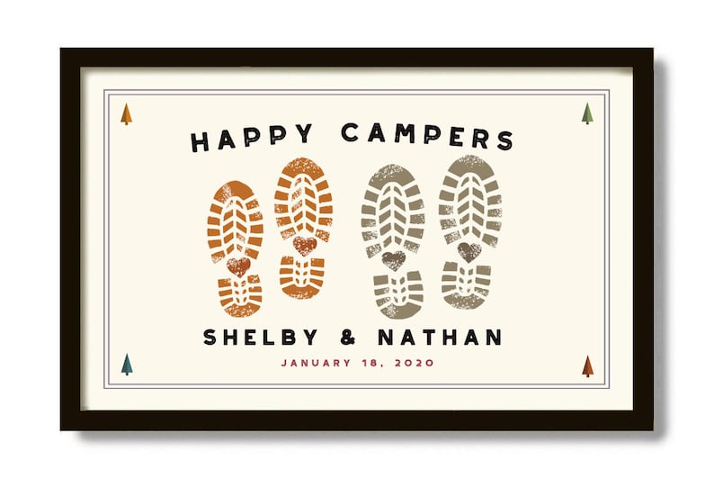 Pode incluir: Impress&atilde;o emoldurada com as palavras "HAPPY CAMPERS" acima de dois pares de pegadas de botas com detalhes de cora&ccedil;&atilde;o. Os nomes "Shelby & Nathan" e a data "18 de janeiro de 2020" est&atilde;o abaixo. A moldura &eacute; marrom escuro.