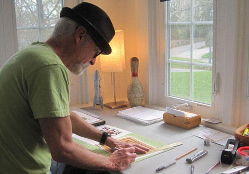 May include: An individual wearing a green t-shirt and a black hat, working at a desk. The desk has various items, including a lamp, a rocket model, a bowling pin, and art supplies. The person is using a ruler and a pen on a drawing.