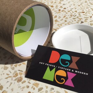 May include: A brown cardboard tube with a green and white logo. The tube is open and a white plastic lid is next to it. A black business card with the text "Dex Mex Art Prints / Vintage & Modern" is in the foreground.