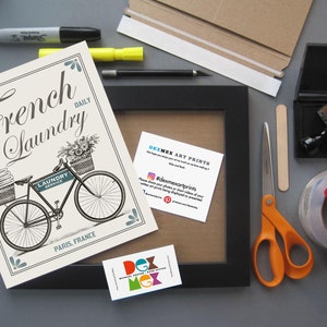 K&ouml;nnte beinhalten: Ein schwarzer gerahmter Druck mit einer Vintage-Illustration eines Fahrrads mit einem Korb voller Blumen und dem Text "French Laundry Daily". Der Druck ist auf wei&szlig;em Hintergrund mit blauen Akzenten. Das Bild zeigt auch verschiedene Materialien zum Einrahmen eines Drucks, wie z. B. ein Lineal, eine Schere, Klebeband und einen Stempel.
