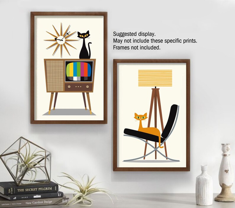 May include: Two framed prints with a mid-century modern theme. The first print features a black cat sitting on top of a vintage television with a clock behind it. The second print features an orange cat sitting in a black chair with a tripod lamp next to it.