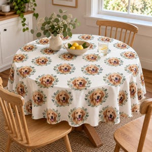 May include: A round, off-white tablecloth featuring a repeating pattern of golden retriever dog faces framed by green eucalyptus-style leaves. The fabric drapes over a circular wooden table, which is surrounded by wooden chairs.