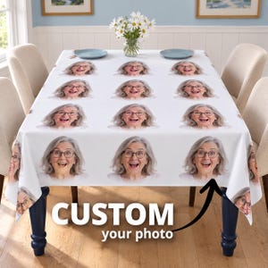 May include: A white tablecloth featuring a repeating pattern of a smiling woman with gray hair and glasses. The tablecloth is draped over a rectangular table with blue legs, and a glass vase with white daisies sits in the center.