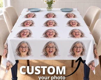 May include: A white tablecloth featuring a repeating pattern of a smiling woman with gray hair and glasses. The tablecloth is draped over a rectangular table with blue legs, and a glass vase with white daisies sits in the center.