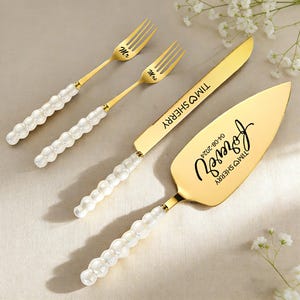 May include: A gold-colored wedding cake serving set with two forks, a knife, and a server. The forks are inscribed with "Mr" and "Mrs". The knife and server are engraved with names and a date. The handles are white with a pearl-like design.