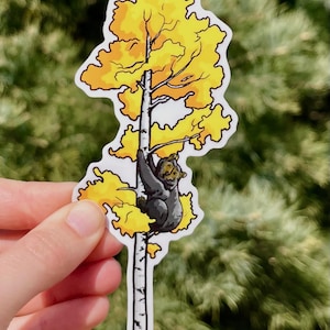 May include: A sticker featuring a cartoon bear climbing a tree with yellow leaves. The bear is gray, and the tree trunk is white with black markings. The background is blurred green.