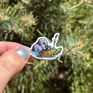 May include: A sticker featuring a cartoon turtle with a bird riding on its back, in a body of water. The turtle has a brown shell and green body. The bird is blue and yellow. A gray rock and cattails are also in the image.