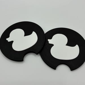 May include: Two black car coasters with a white duck silhouette design. The coasters have a circular shape with a small notch on one side. The duck design is a simple, clean outline.