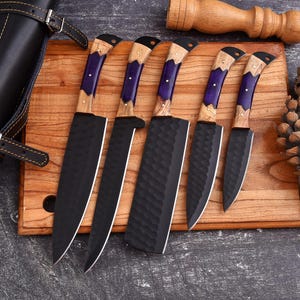 Op de afbeelding: Een set van vijf zwarte keukenmessen met houten en paarse handvatten, gerangschikt op een houten snijplank. De messen hebben een getextureerde afwerking. Een zwarte leren schede is links zichtbaar. Een houten hamer staat op de achtergrond.