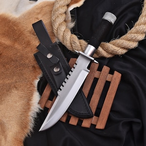 May include: A large survival knife with a serrated blade and a black wrapped handle. The knife is in a black leather sheath and rests on a wooden stand. A thick rope and animal fur are in the background.
