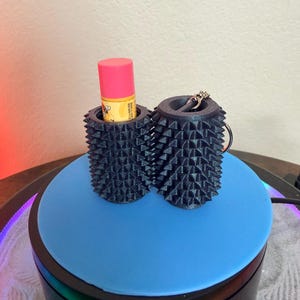 May include: Two cylindrical black holders with a textured, spiked exterior surface. One holder contains a pink-capped lip balm tube, while the other features a metal keychain ring attached to the top edge, providing a secure way to carry small items.