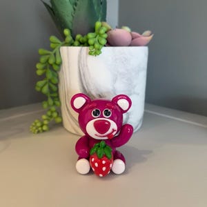 Könnte beinhalten: Eine magenta Bärenfigur mit weißen Akzenten, die eine rote Erdbeere mit grünen Blättern hält. Der Bär hat einen freundlichen Ausdruck. Die Figur steht auf einer hellgrauen Oberfläche, mit einem dekorativen Pflanzgefäß im Hintergrund.