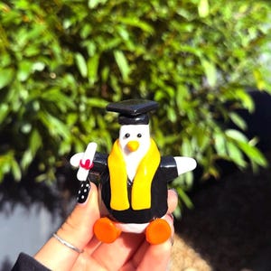 May include: A small penguin figurine wearing a black graduation cap and gown with a yellow stole. The penguin has a white body, orange feet, and a small orange beak. It holds a white rolled diploma tied with a red ribbon in one wing.