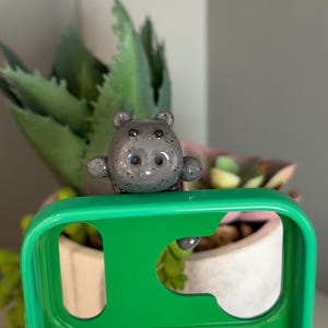 May include: A small, grey, speckled hippo figurine attached to the top edge of a bright green phone case. The hippo has a smooth, glossy finish with dark grey spots, small rounded ears, and a wide snout, positioned over the camera cutout area.