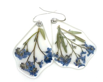 Puede incluir: Un par de pendientes de resina transparente con un diseño floral. Cada pendiente presenta nomeolvides azules y hojas verdes encapsuladas en una resina transparente en forma de ala. Los pendientes tienen ganchos plateados.