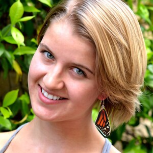 May include: A woman with blonde hair wearing a gray tank top and butterfly earrings. The earrings are silver with a brown and orange butterfly design.