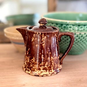 May include: A small, brown ceramic pitcher with a lid and handle. The pitcher has a speckled, textured pattern in shades of brown and tan. The background includes other ceramic bowls.