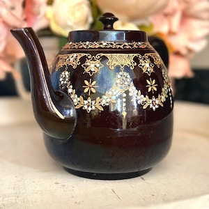 May include: A dark brown ceramic teapot with a lid and a curved spout. The teapot is decorated with gold floral patterns and small, light blue accents. The background features blurred pink and white flowers.