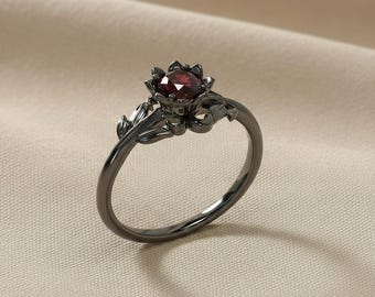 May include: A dark gray ring with a flower-shaped setting holding a round, deep red gemstone. The ring's band features delicate floral and leaf details, creating an elegant and nature-inspired design. The ring is set against a neutral background.