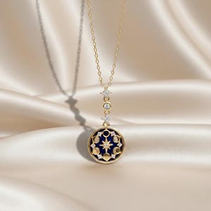 Puede incluir: Un collar dorado con un colgante celestial. El colgante presenta un fondo de esmalte azul oscuro con un dise&ntilde;o de estrellas y fases lunares. El collar tiene una cadena delicada y est&aacute; adornado con peque&ntilde;os detalles brillantes.