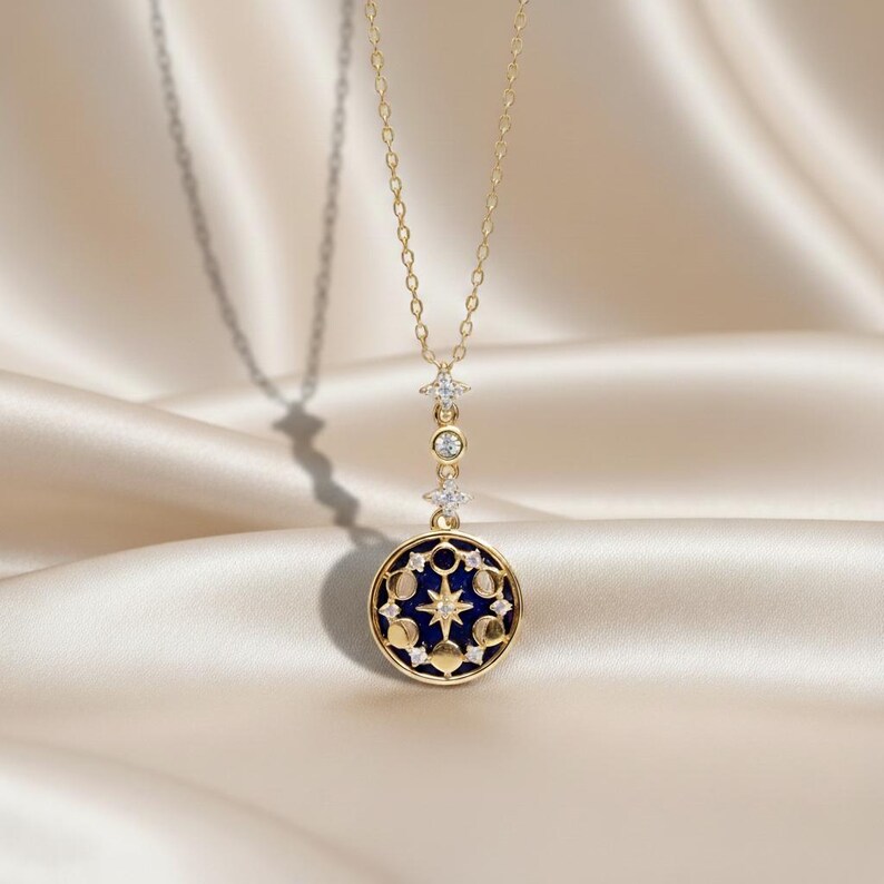 Puede incluir: Un collar dorado con un colgante celestial. El colgante presenta un fondo de esmalte azul oscuro con un dise&ntilde;o de estrellas y fases lunares. El collar tiene una cadena delicada y est&aacute; adornado con peque&ntilde;os detalles brillantes.