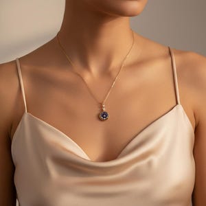 Puede incluir: Un collar dorado con un colgante circular. El colgante tiene una piedra central azul oscuro rodeada de piedras m&aacute;s peque&ntilde;as. El collar lo lleva una persona que lleva una blusa de sat&eacute;n.